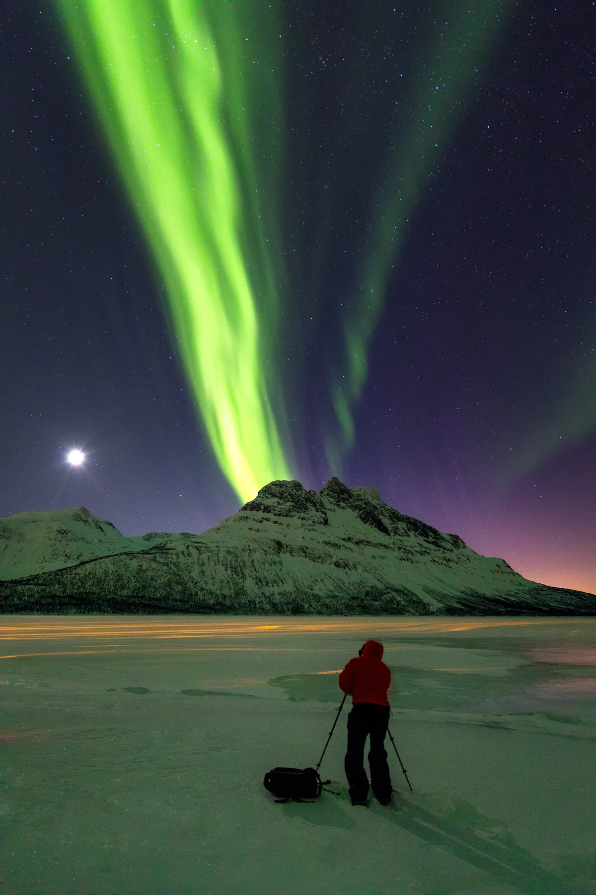 Person unter dem Nordlicht mit Mond über dem Nova – Lofoten, Norwegen